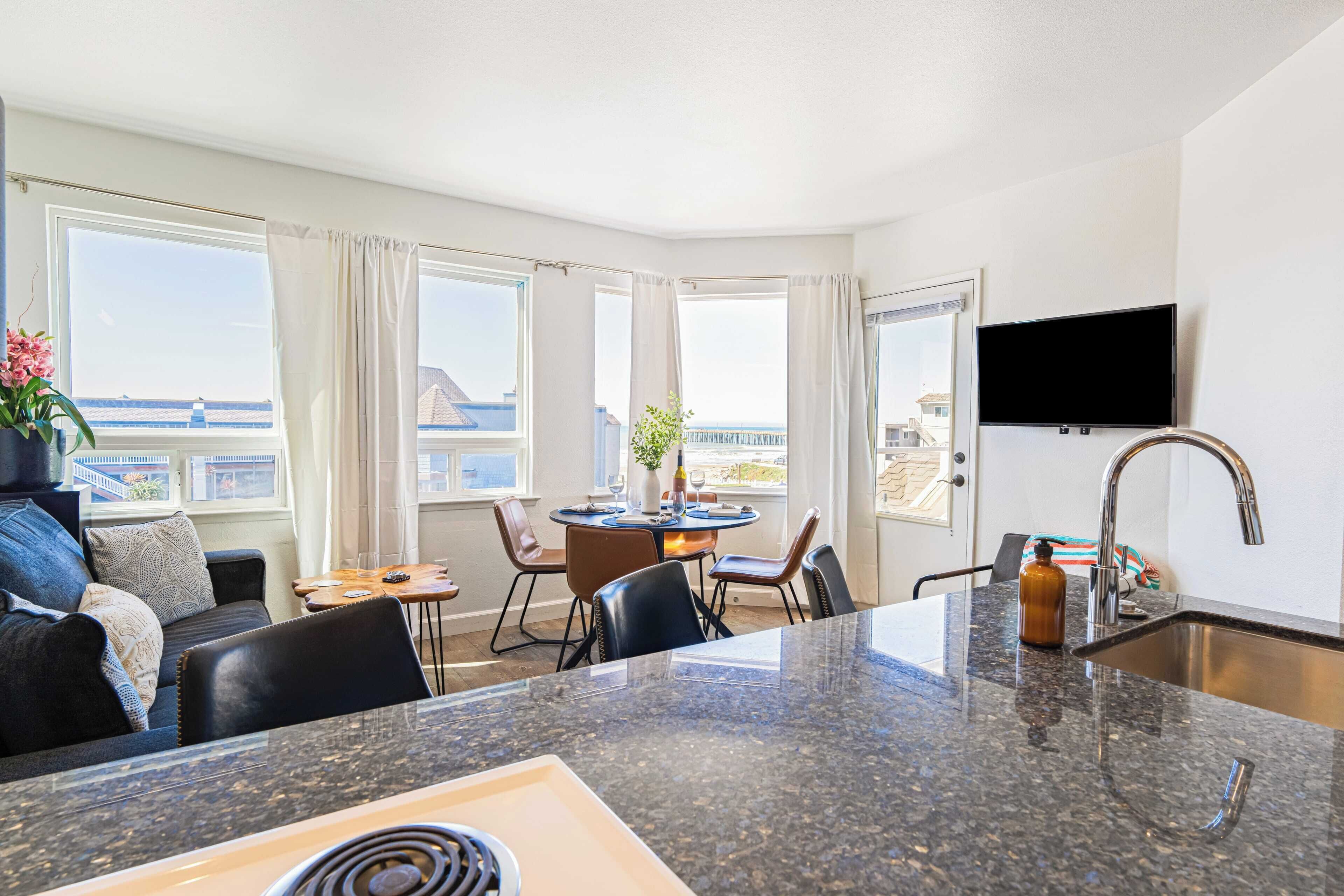 A modern kitchen with a granite countertop, a dining area with a round table and chairs, a sofa, and a TV mounted on the wall.