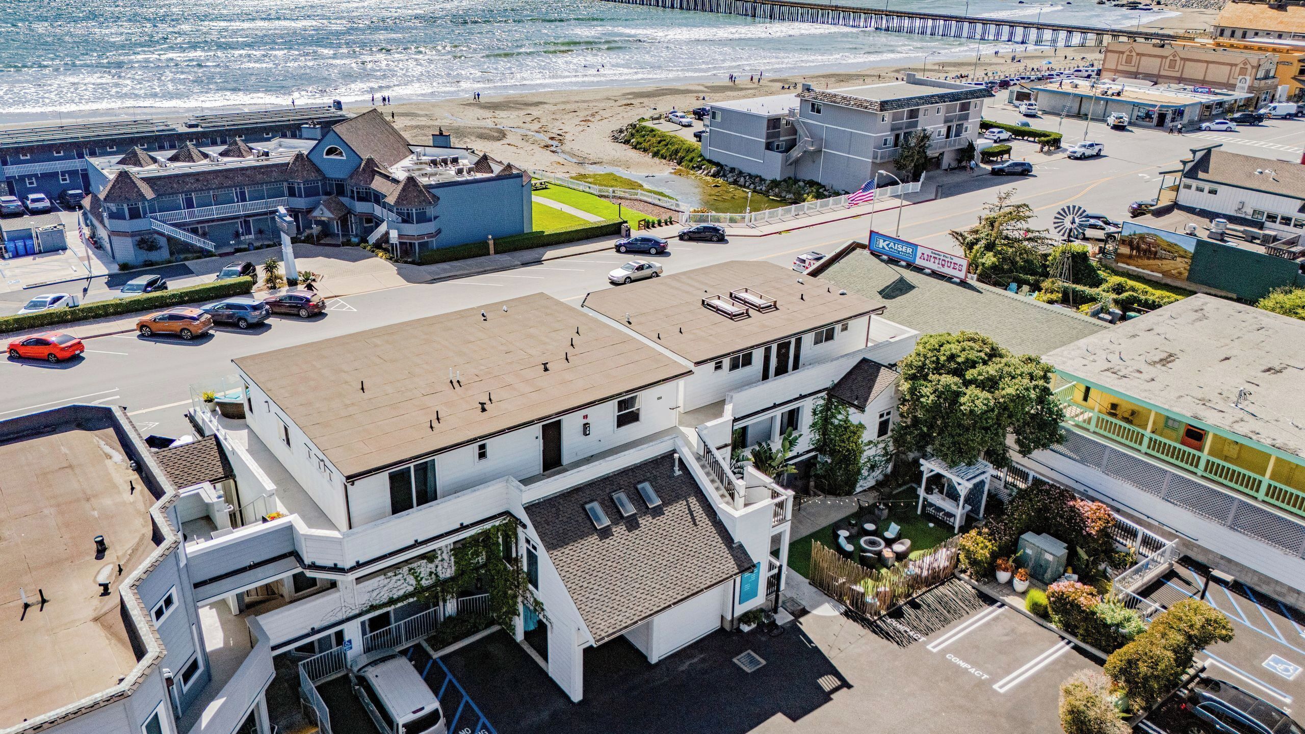 An aerial view of a coastal town with a prominent hotel featuring a rooftop terrace, a parking area, and a beachfront promenade.