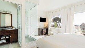 Habitación de hotel con cama, baño con ducha de vidrio, televisor y vistas al exterior.