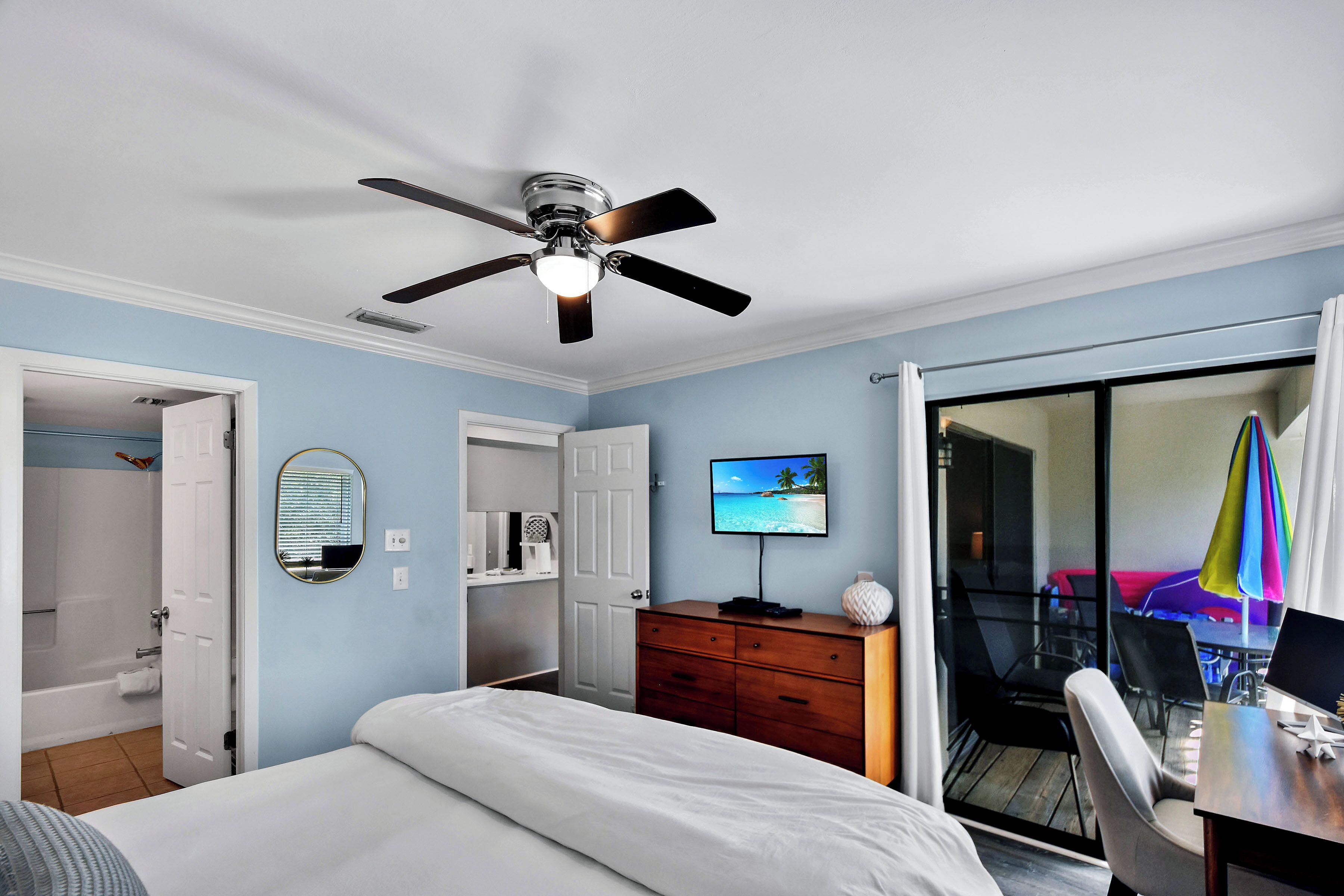 Un dormitorio con cama, ventilador de techo, televisor, cómoda y puerta corrediza de vidrio que da a un balcón con mesa y sillas.