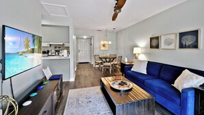 A modern living room with a blue sofa, a coffee table, and a flat-screen TV displaying a beach scene. - Oceanfront Condo - Private Boardwalk - Pool & Balcony (St. Simons Island)