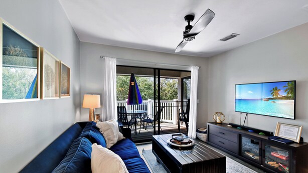 A modern living room with a dark blue sofa, a coffee table, a flat-screen TV, and a balcony with outdoor furniture. - Oceanfront Condo - Private Boardwalk - Pool & Balcony (St. Simons Island)