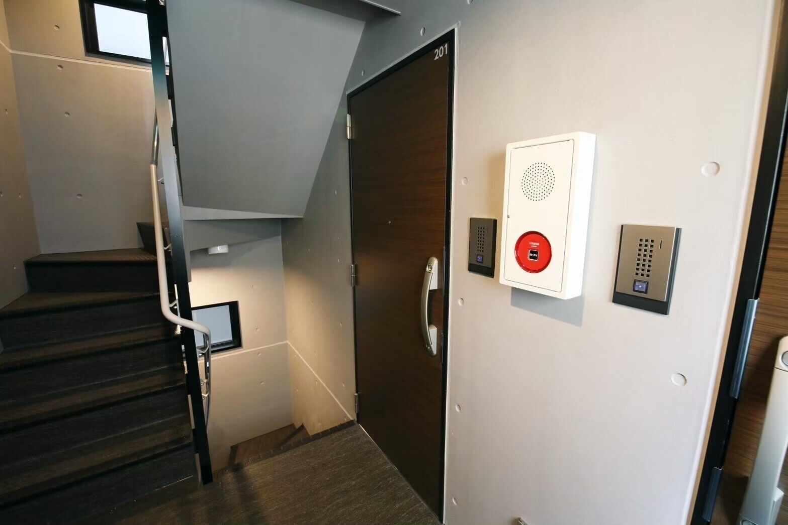A modern staircase with a handrail, a door with a number plate, and a wall-mounted intercom system.
