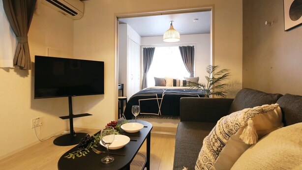 Een moderne woonkamer met een flatscreen televisie, een eettafel met borden en wijnglazen, een bank, en een uitzicht op de slaapkamer met een bed en gordijnen.