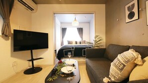 Een moderne woonkamer met een flatscreen-tv, een bank, een salontafel met een wijnglas en druiven, en uitzicht op een slaapkamer met een bed en gordijnen.