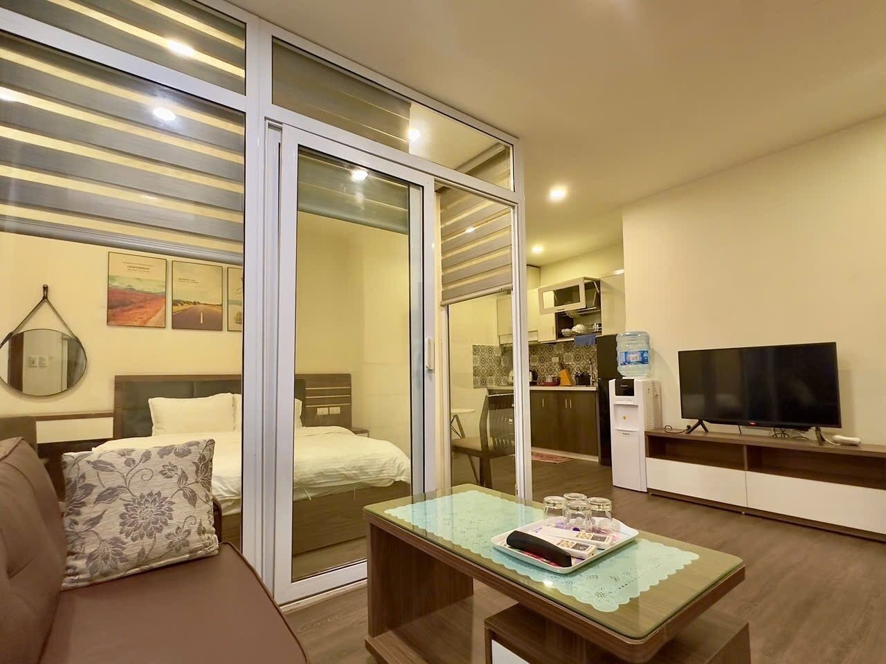 A hotel room with a bed, a sofa, a dining table, a TV, and a sliding glass door leading to a kitchen area.