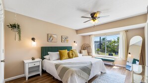 A bedroom with a bed, bedside tables, a ceiling fan, a window with curtains, and a view of greenery outside.