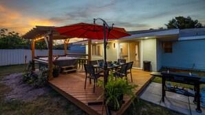 A backyard deck with a dining table, chairs, a red umbrella, and a hot tub.