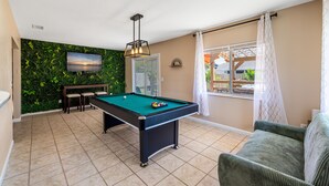 A pool table with chairs, a television on a green wall, and a view of a house through a window.