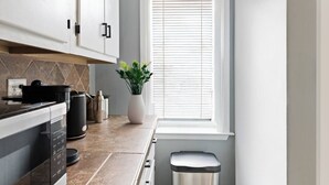 Une cuisine moderne avec des meubles blancs, un plan de travail sur lequel se trouve une machine à café, une plante et une fenêtre avec des stores.