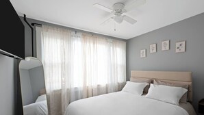 Une chambre à coucher avec un lit, un ventilateur de plafond, une fenêtre avec des rideaux légers et des tableaux encadrés accrochés au mur.