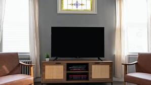 Un salon équipé d’un téléviseur à écran plat, de deux fauteuils en cuir, d’un meuble de rangement en bois où sont posés des livres, et d’une petite plante.