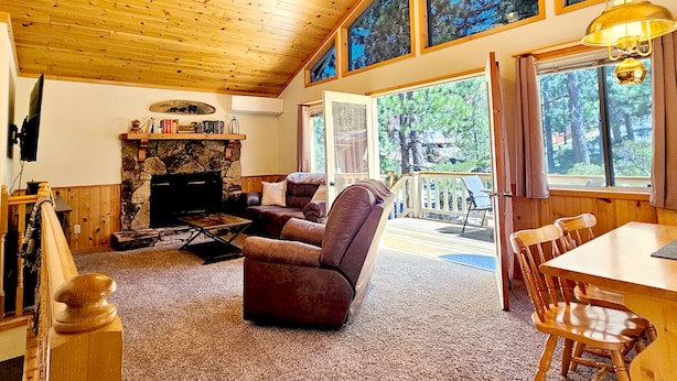 A cozy living room with a stone fireplace, wooden ceiling, and large windows offering a view of trees.