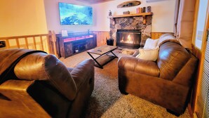 A cozy living room with leather sofas, a stone fireplace, a flat-screen TV, and a wooden ceiling.