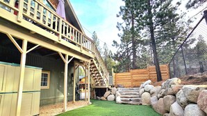 A rustic cabin with a wooden deck, a staircase leading to a balcony, and a stone wall.