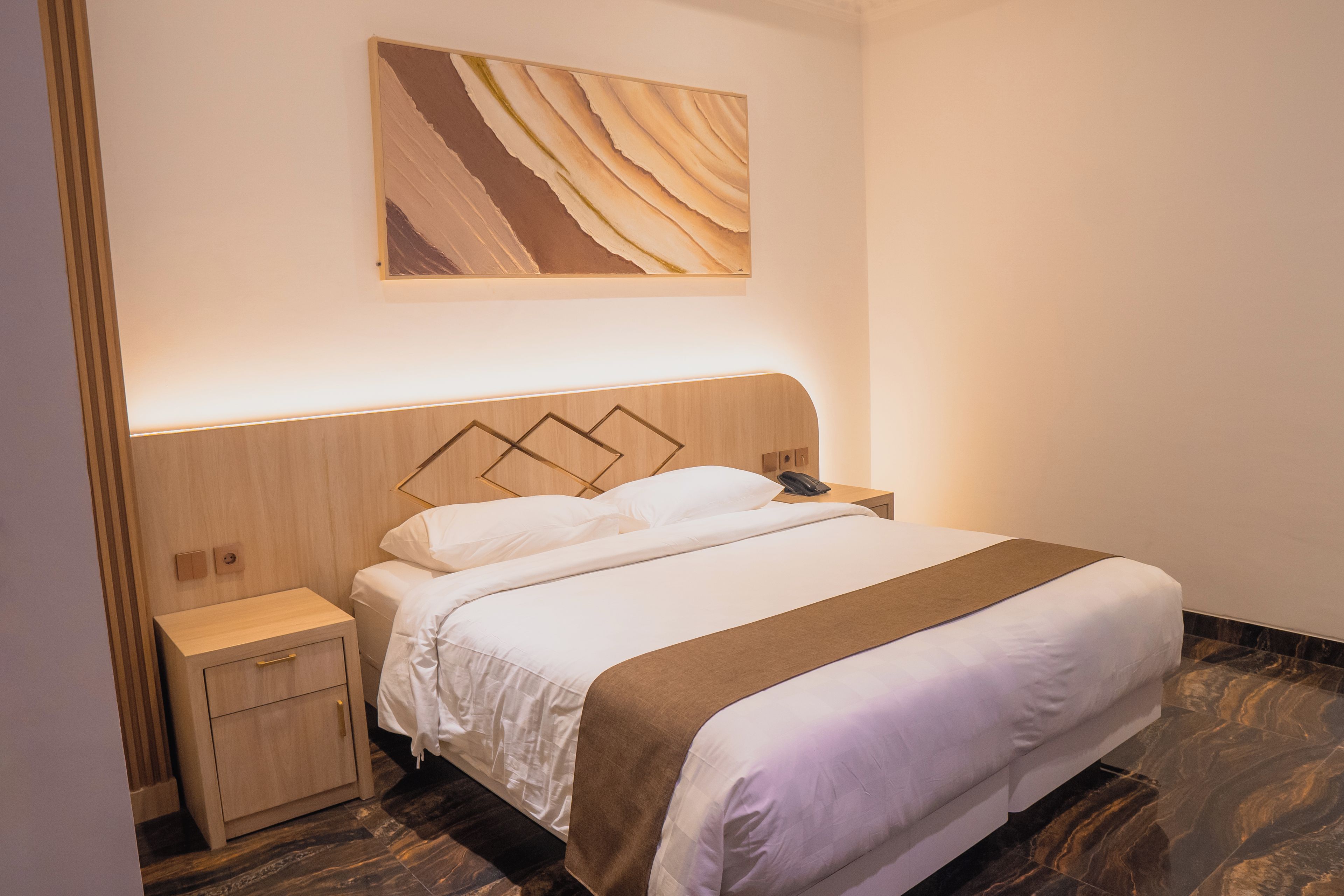 Quarto de hotel com cabeceira de madeira, uma cama grande com roupa de cama branca e manta marrom, mesa de cabeceira com abajur e uma obra de arte pendurada na parede.