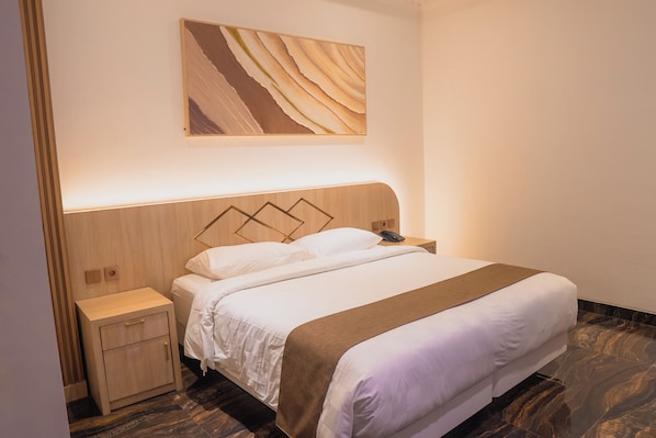Quarto de hotel com cabeceira de madeira, uma cama grande com roupa de cama branca e manta marrom, mesa de cabeceira com abajur e uma obra de arte pendurada na parede.