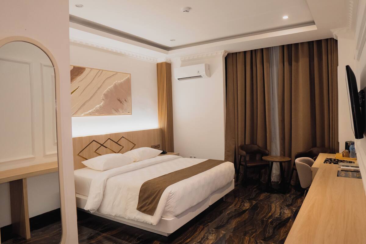 A hotel room with a bed, a desk, a chair, a TV, and an air conditioning unit.