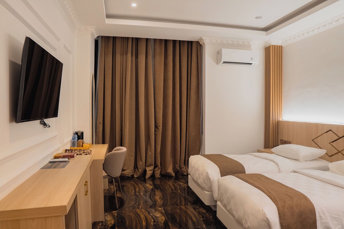 A hotel room with a desk, chair, two beds, a flat-screen TV, and a large window with curtains.