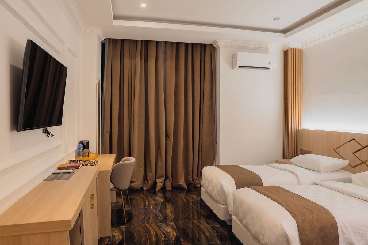 A hotel room with a desk, chair, two beds, a flat-screen TV, and a large window with curtains.