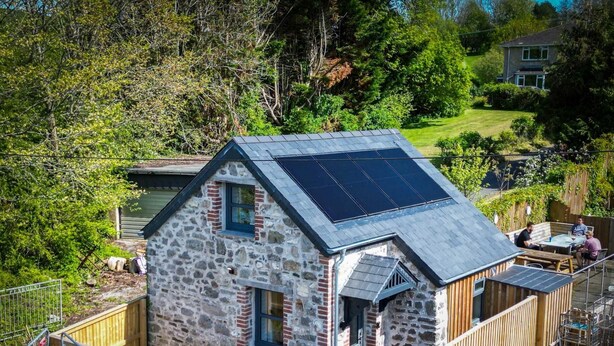 Un edificio de piedra con techo inclinado y paneles solares, rodeado por una cerca de madera y follaje.