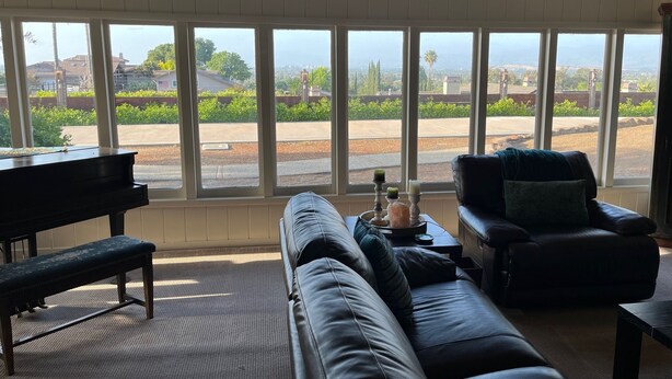 A living room with a piano, a couch, and a chair, with large windows offering a view of a residential area.