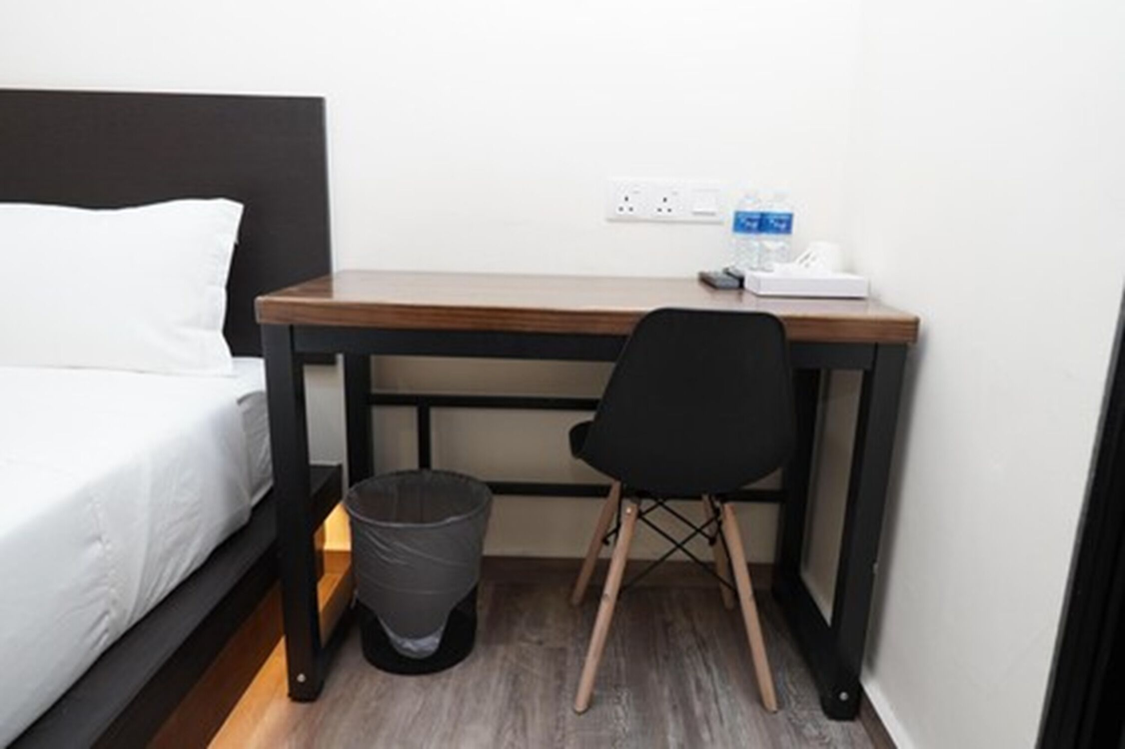 A compact hotel room with a bed, a desk, a chair, a trash bin, and a water bottle on the desk.