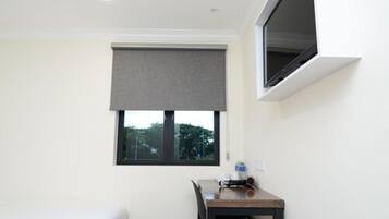 A hotel room with a bed, a small table with a chair, a window with a blind, and a TV mounted on the wall.
