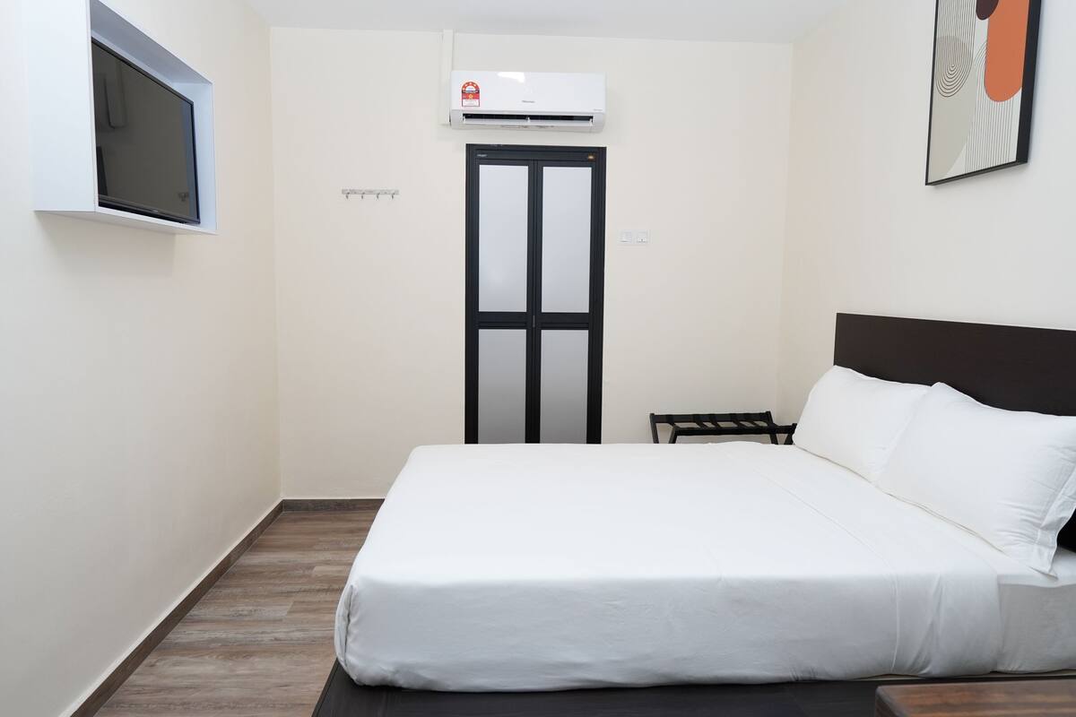 A hotel room with a bed, a small table, a TV mounted on the wall, and an air conditioning unit.