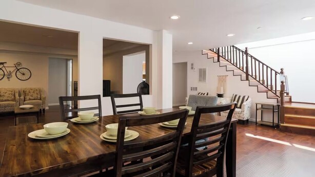 A dining area with a wooden table set for four, surrounded by dark wooden chairs. A staircase with a wooden railing leads to an upper level.