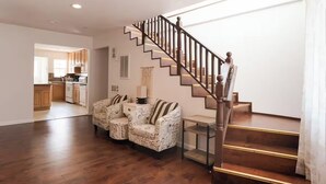 A spacious living area with a wooden staircase, a kitchen in the background, and patterned armchairs.