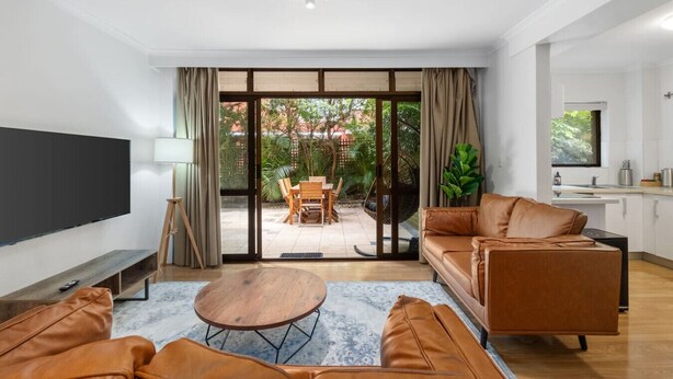 Ein modernes Wohnzimmer mit großem Fernseher, Ledersofas, einem Couchtisch und einer Glastür, die auf eine Terrasse mit Essplatz führt.