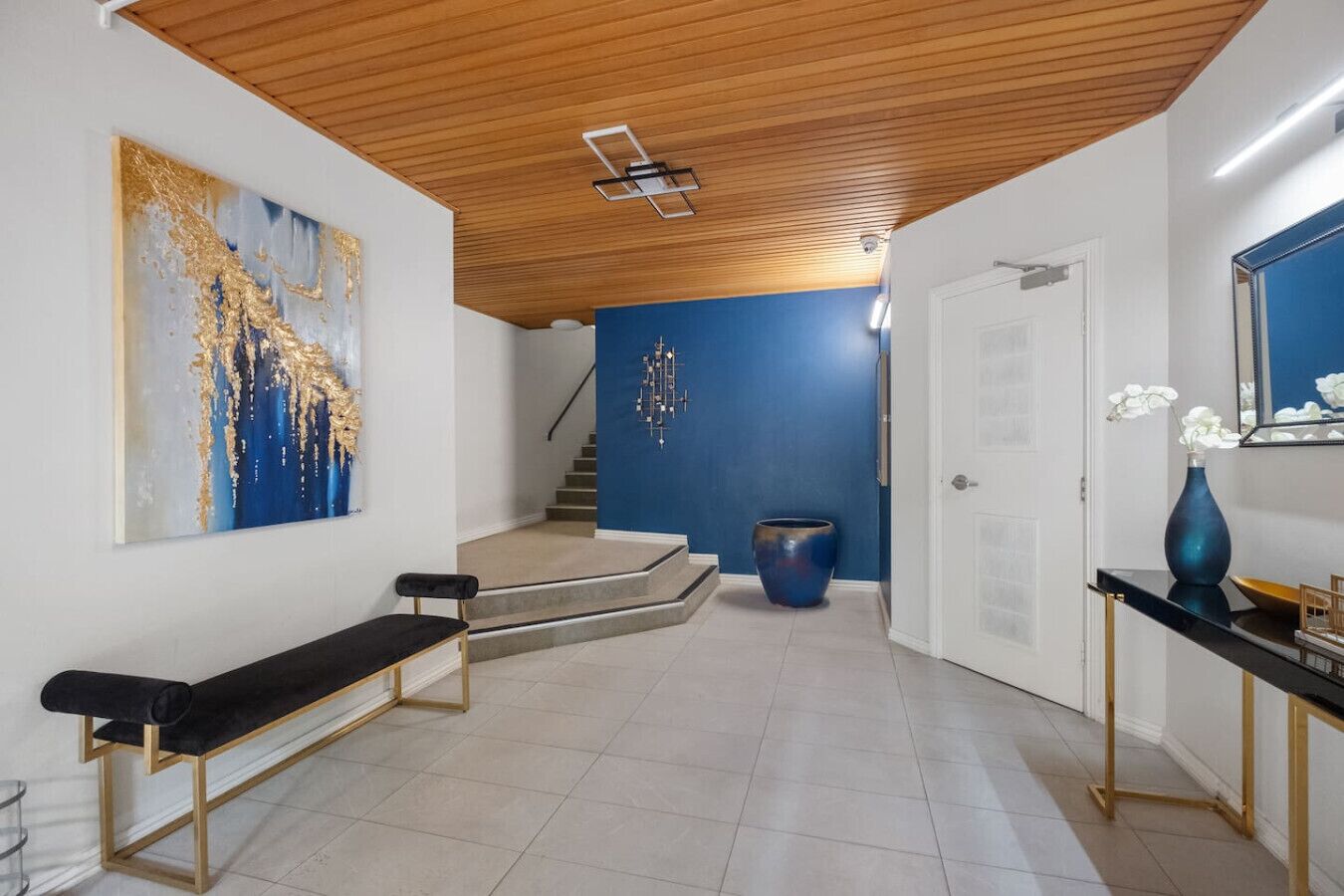 A modern interior with a wooden ceiling, a blue accent wall, a large abstract painting, a bench, a vase, and a staircase.