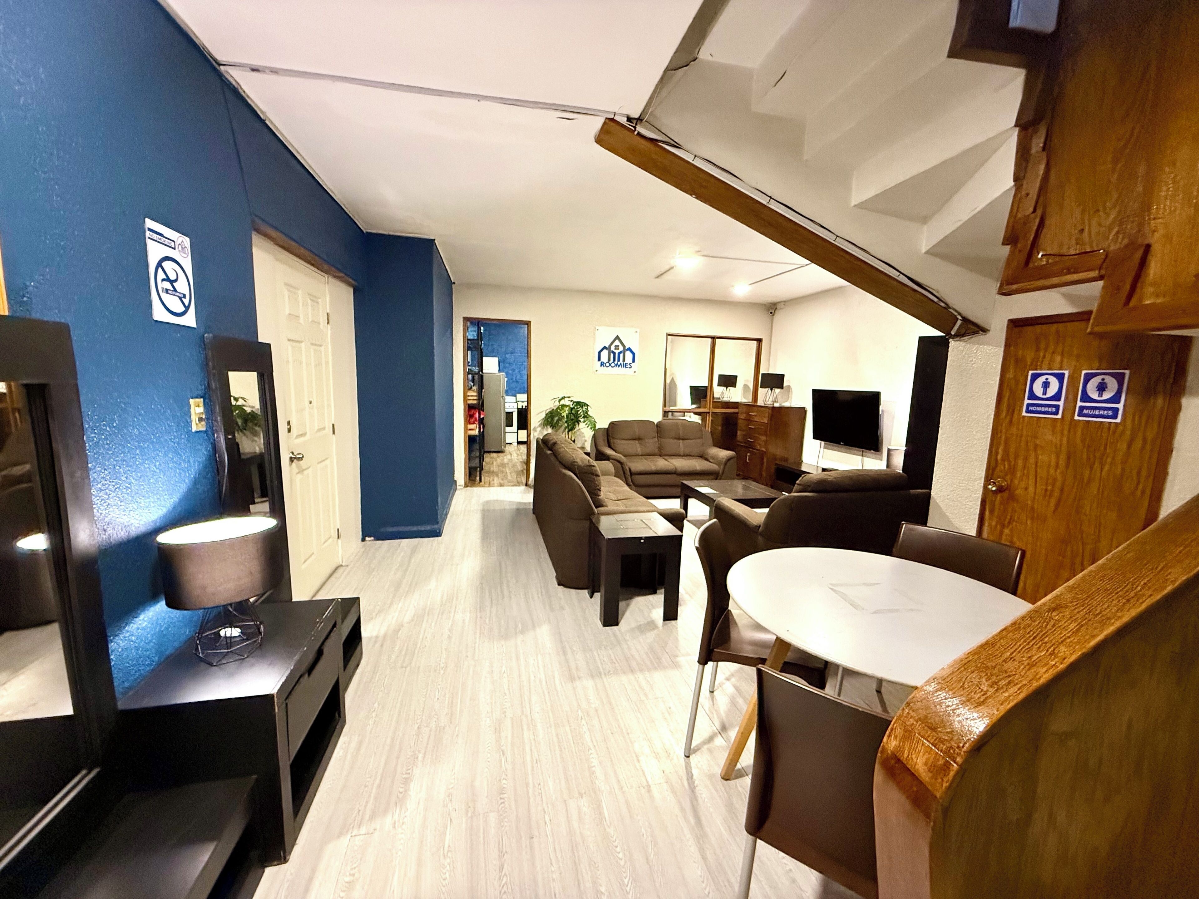 A modern living room with a sofa set, a coffee table, and a TV. There is a dining table with chairs. The room has a blue accent wall and wooden flooring.