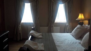 A hotel room with a bed, two windows with curtains, a radiator, a bedside lamp, and a dresser.