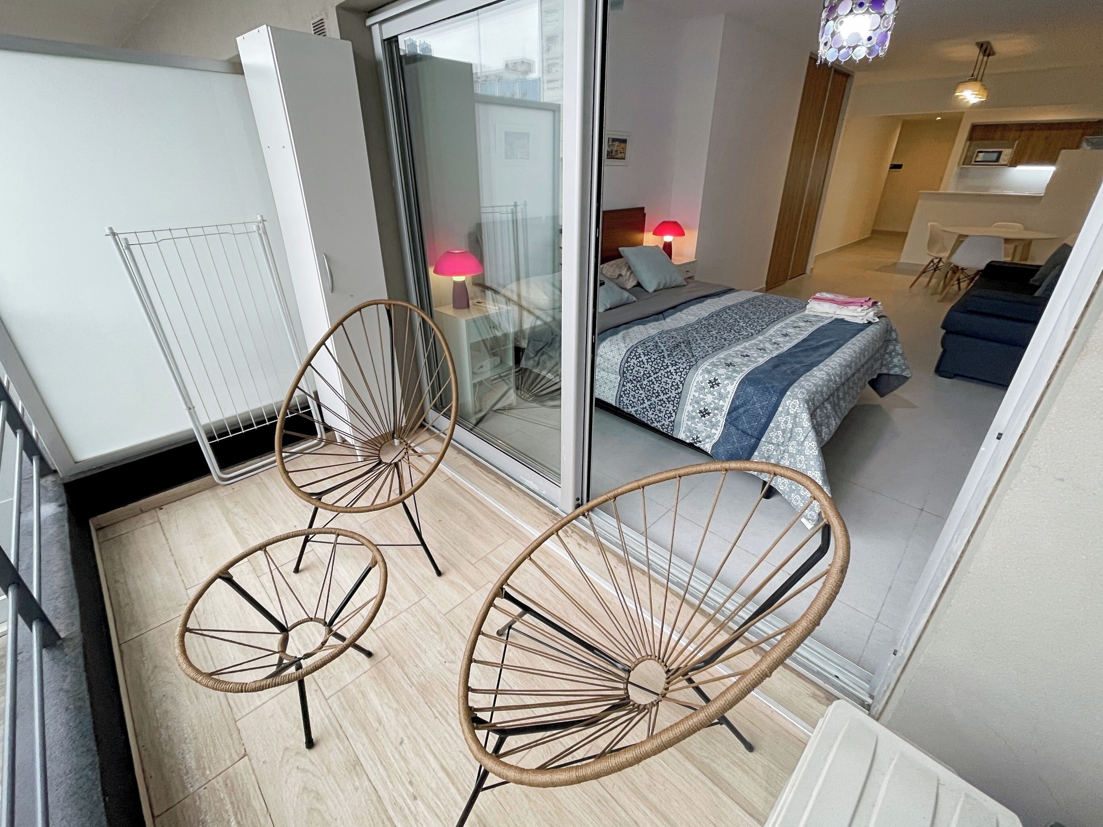 Une chambre moderne avec une porte vitrée donnant sur un balcon, un lit avec une literie à motifs et deux fauteuils en osier sur le balcon.