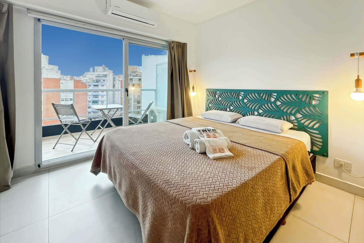 A hotel room with a bed, a balcony with a chair, and a city view.