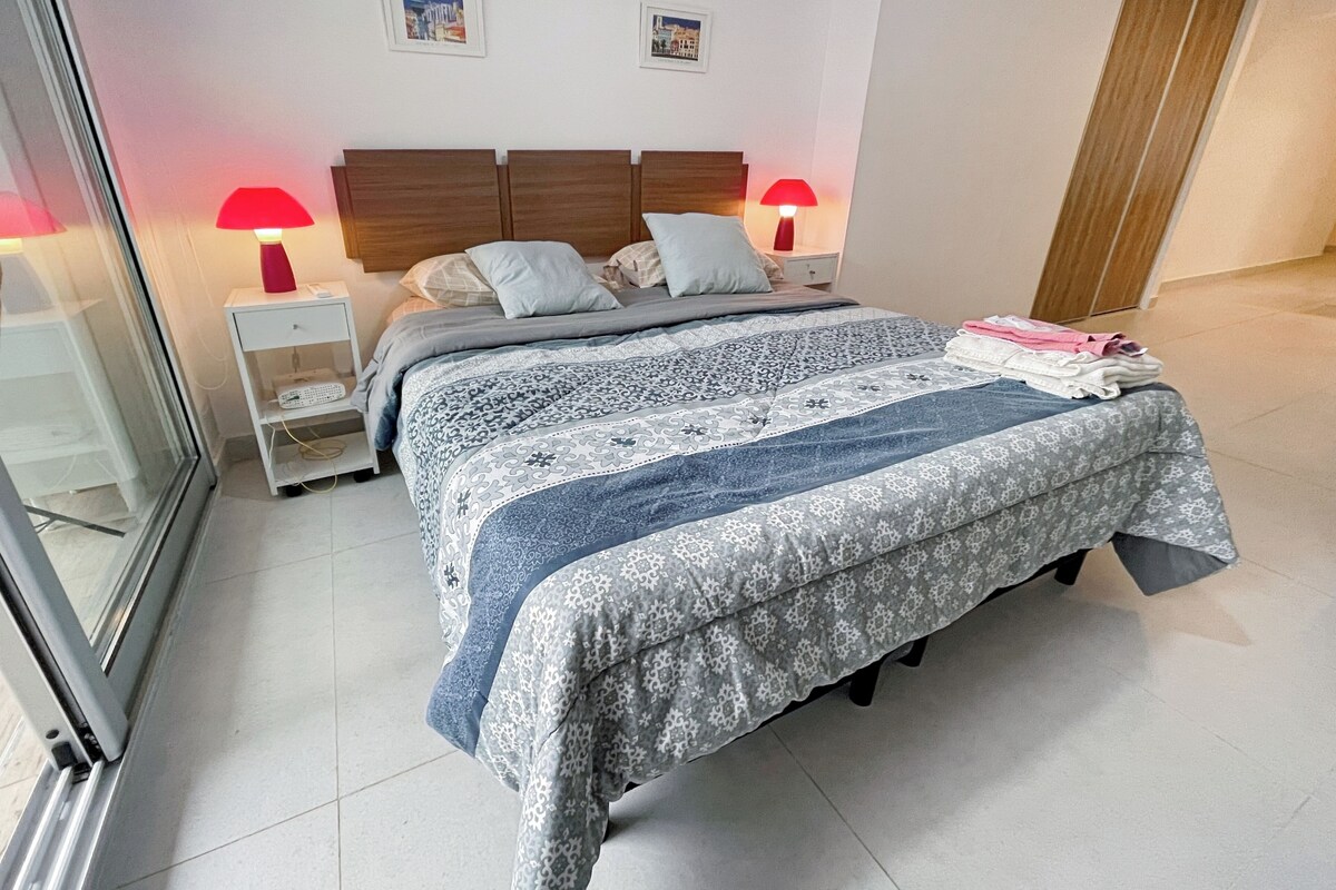 A neatly made bed with patterned bedding, a bedside table with a lamp, and a small shelf.