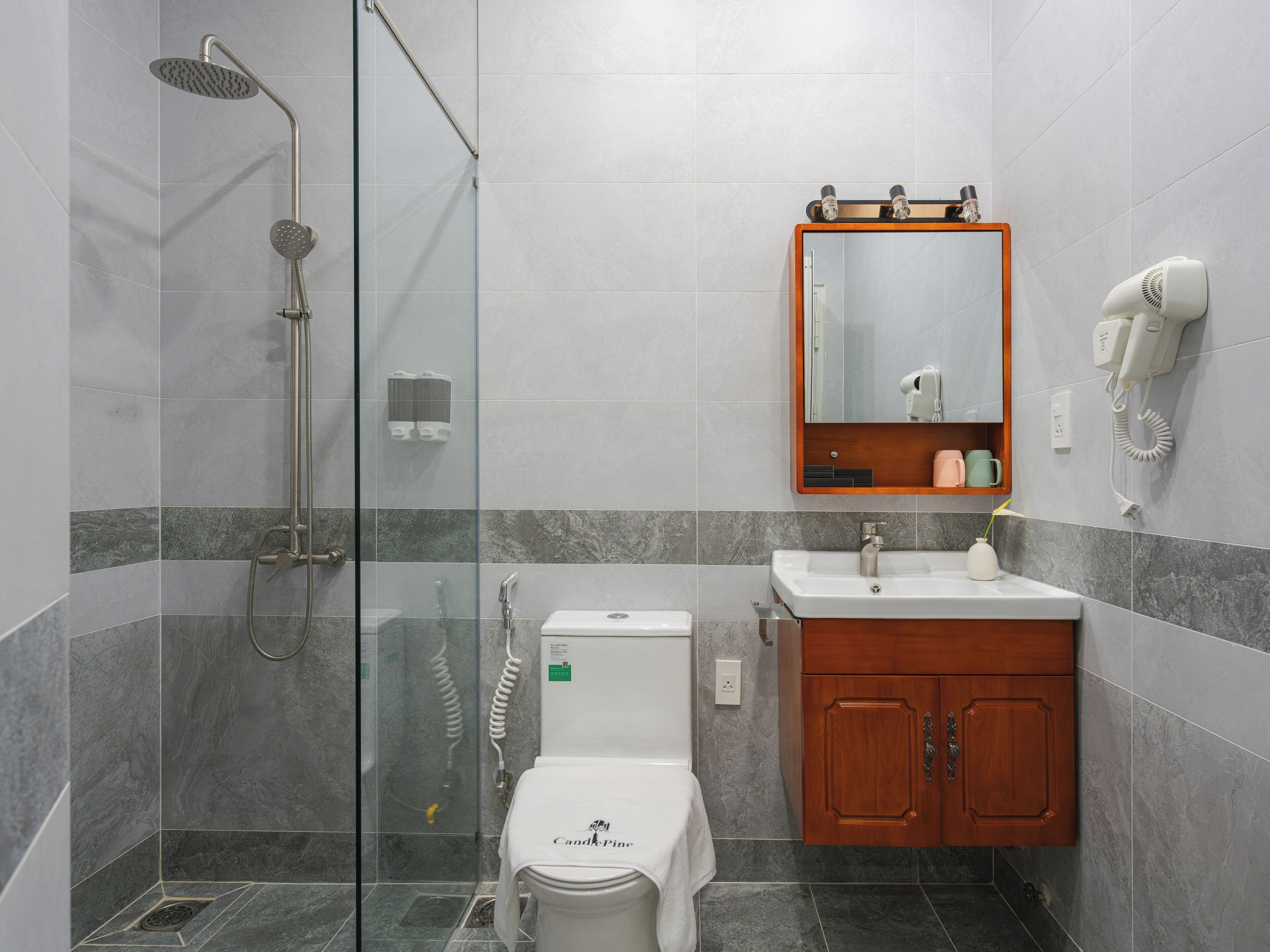 A modern bathroom with a glass-enclosed shower, a white toilet, a wooden vanity with a mirror, and a hairdryer mounted on the wall.