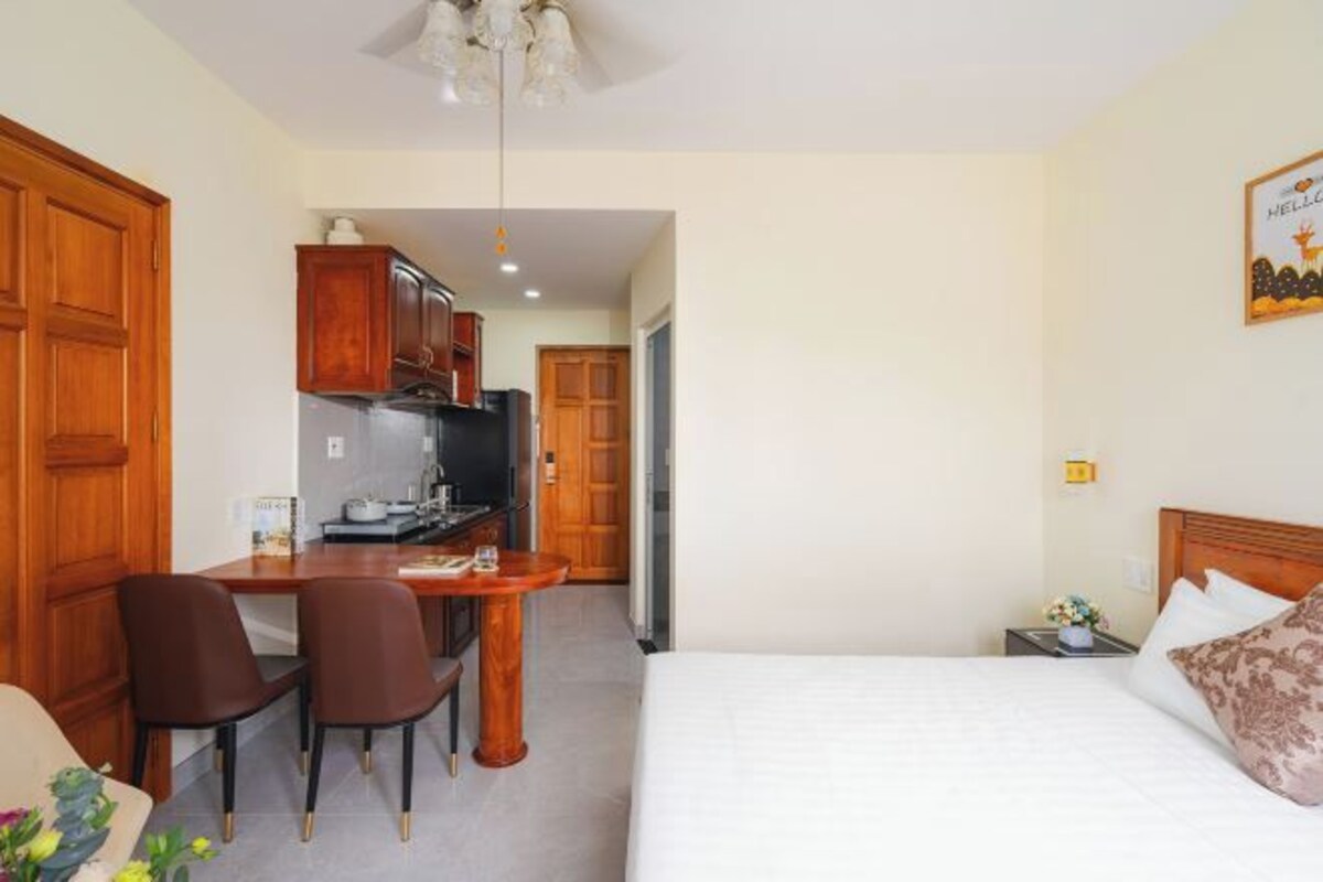 A compact hotel room with a bed, a small desk with chairs, a kitchenette, and a wooden wardrobe.