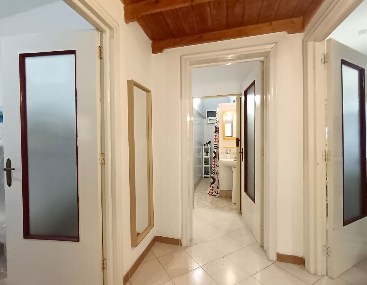 A hallway with white walls and a wooden ceiling. There are doors on both sides, leading to other rooms. The floor is tiled, and a bathroom with a shower and sink is visible in the background.