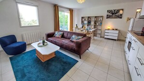 Un salon moderne avec un canapé en cuir marron, un fauteuil bleu et une table basse blanche. On y trouve également un coin repas avec une table et des chaises, ainsi qu'une cuisine avec des meubles blancs et une cuisinière.
