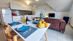 Une cuisine moderne avec des meubles blancs, une table à manger dressée avec des sets de table bleus, des assiettes et des couverts, et un saladier rempli de bananes et d'oranges.