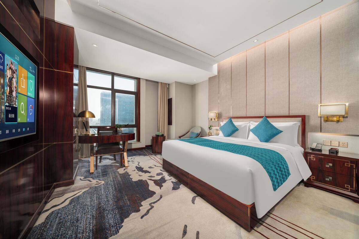 A modern hotel room with a large bed, a desk, and a TV on the left wall.