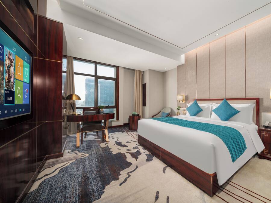 A modern hotel room with a large bed, a desk, and a TV on the left wall.
