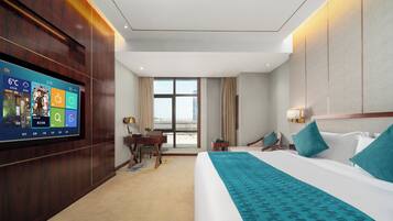 Habitación de hotel con un televisor de pantalla plana grande en la pared, una cama con una manta color turquesa, un escritorio con una silla y vistas a la ciudad desde la ventana.