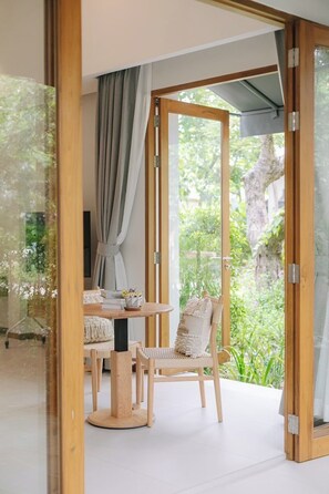 A room with a glass door leading to a garden, a round wooden table with chairs, and a view of greenery outside.