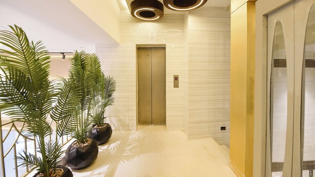 A modern hallway with a marble floor, a glass elevator, and potted plants.