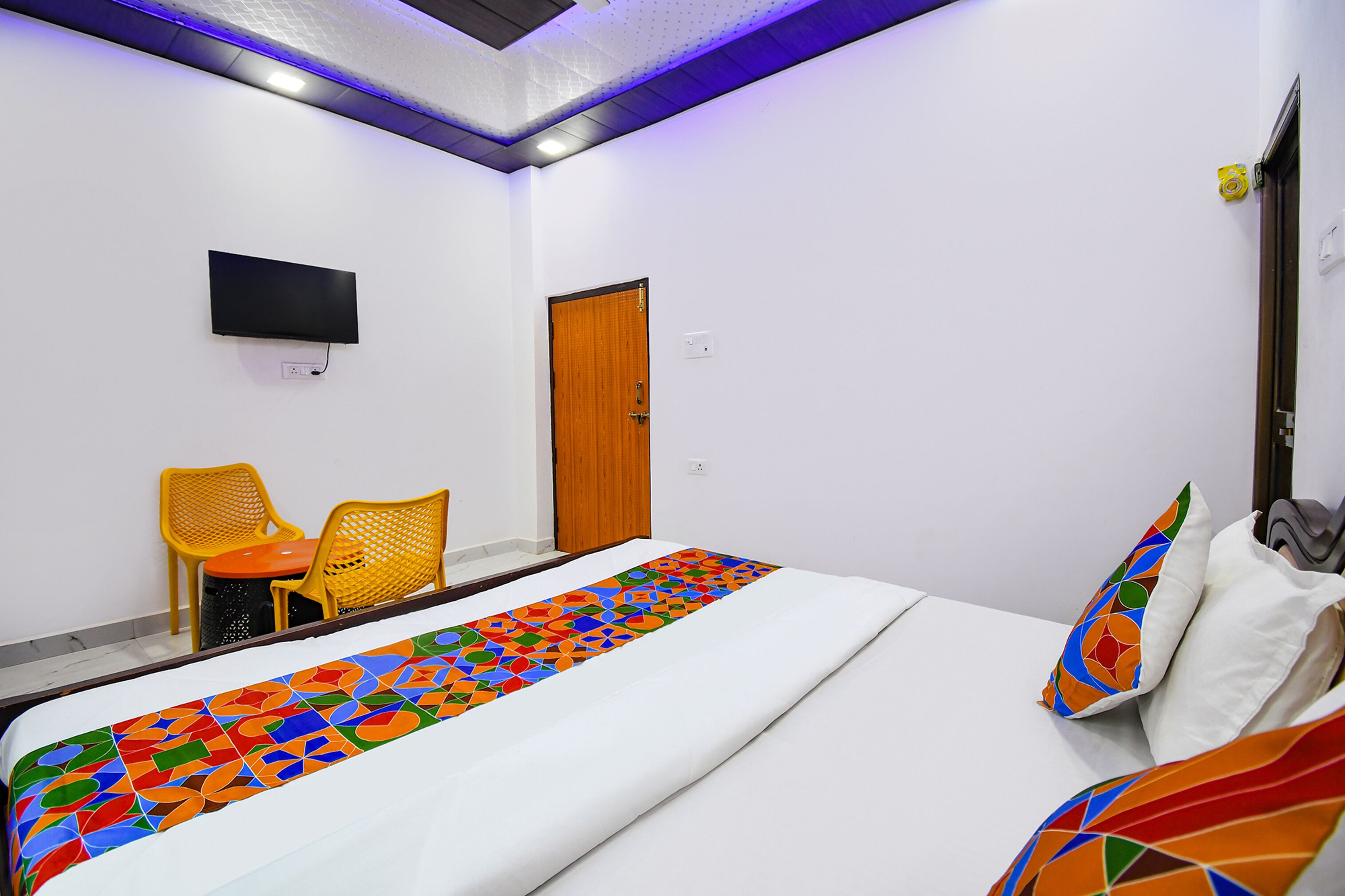 Une chambre d’hôtel avec un lit, un téléviseur fixé au mur, deux chaises jaunes et une porte en bois.