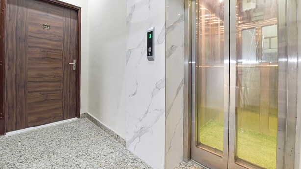 Un couloir avec une porte en bois, un mur en marbre et un ascenseur doté d’un indicateur lumineux vert.